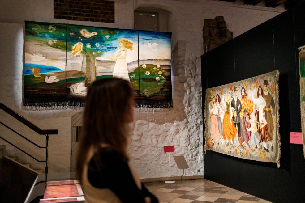 Two large textile artworks are displayed against a stone wall in a museum: one shows a woman and birds, and the other a group of people in folk costumes, with a blurred visitor in the foreground.