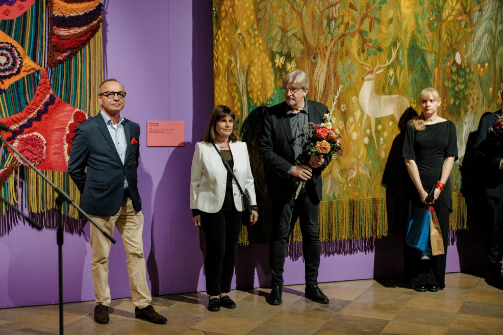 Four people stand in a museum gallery beside a purple wall with textile and tapestry works, including a large forest scene tapestry with deer.