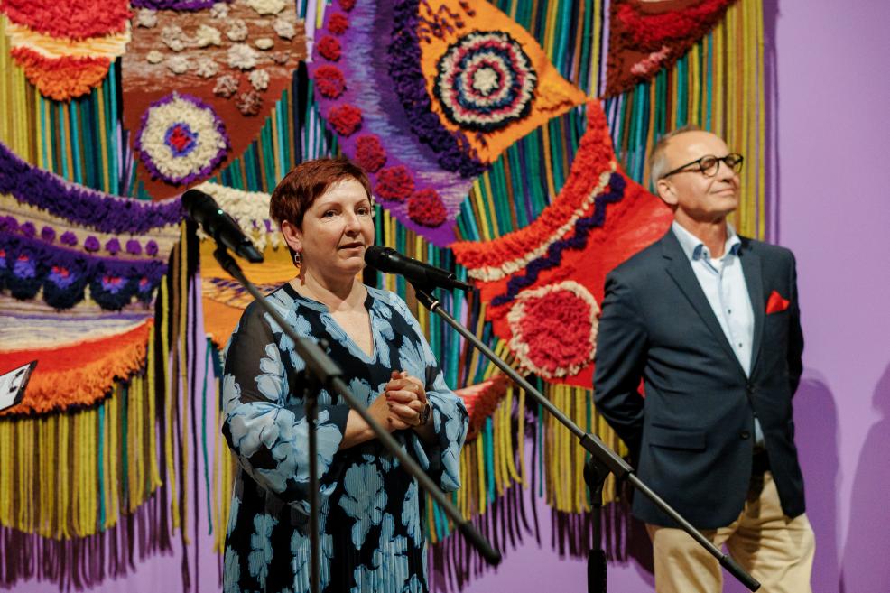 Two people stand at microphones in front of a colorful textile installation in a museum.