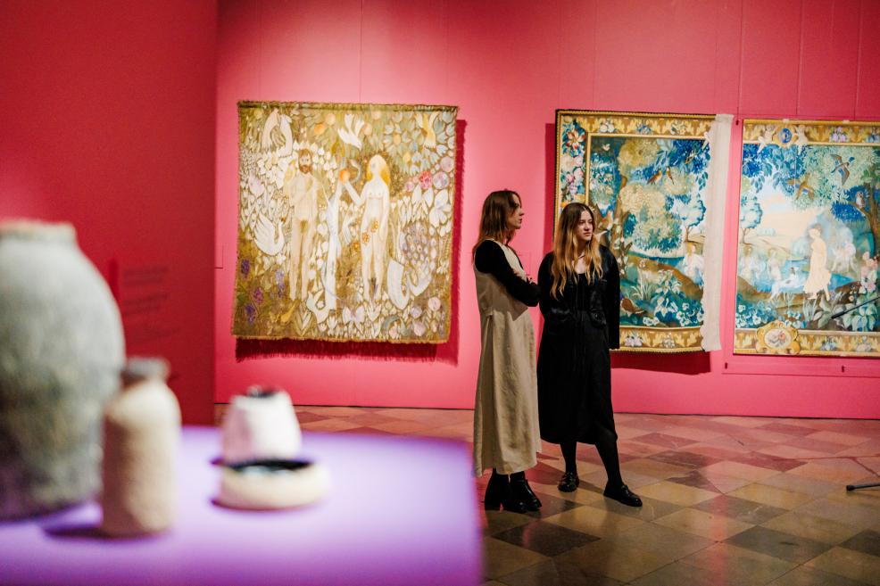Two women stand in a museum gallery with pink walls beside three large textile works with figurative and botanical compositions.