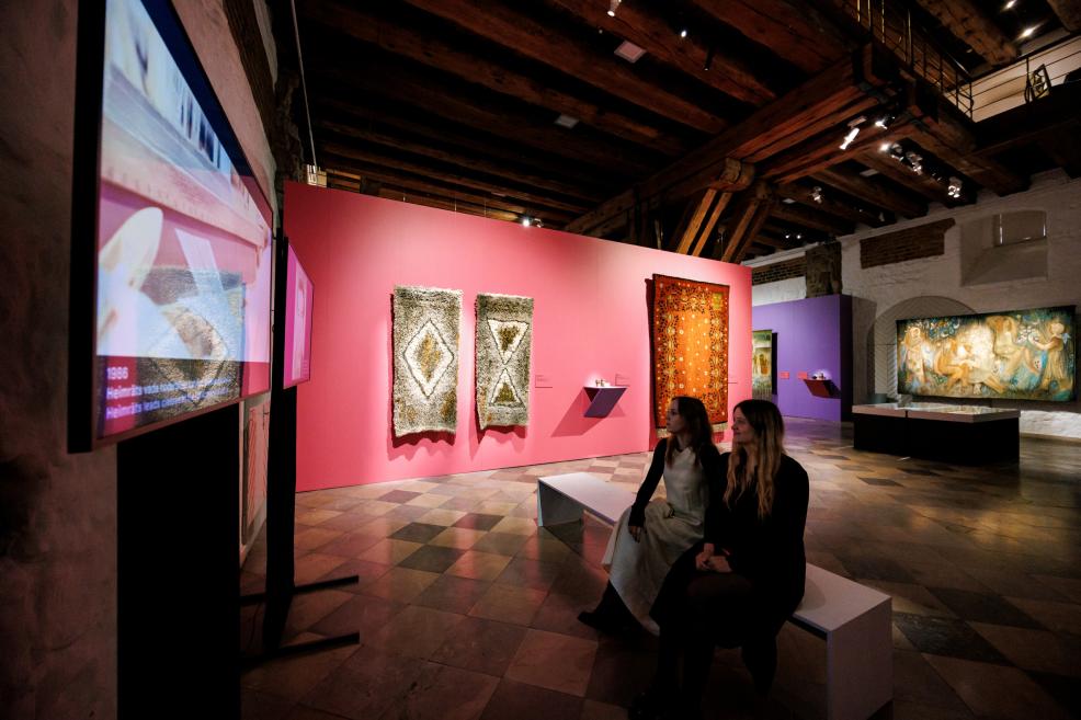 In a museum exhibition hall with pink and purple walls, wooden beams and tiled floor, various artworks are on display, including a video screen, textile pieces and a painting, while two women sit on a bench in the foreground.