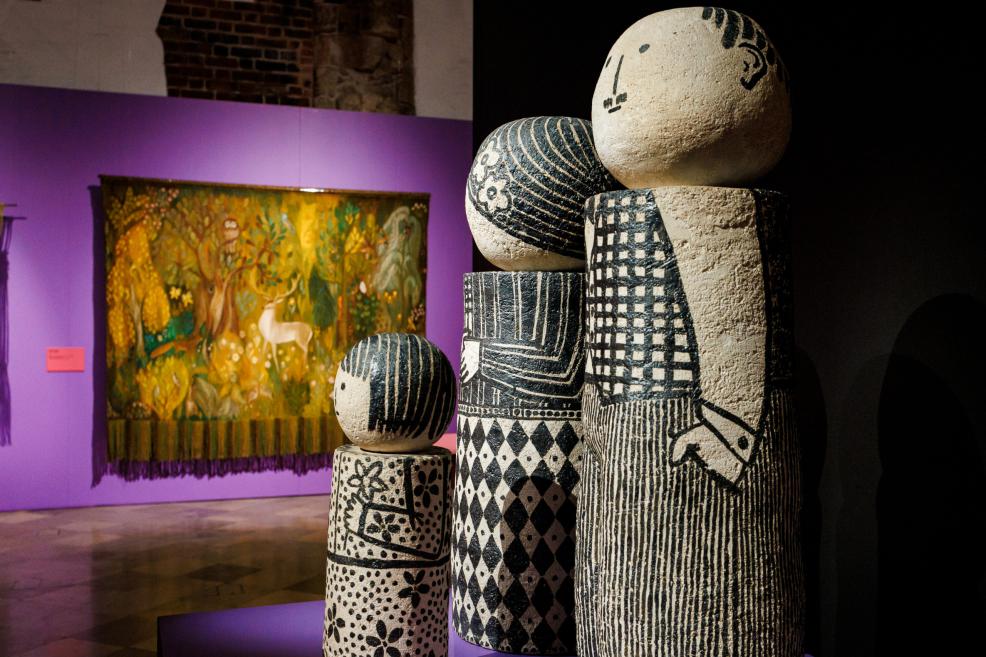 Four decorative cylindrical sculptures with black-and-white patterns stand in an exhibition hall, with a colorful forest-motif tapestry hanging on a purple wall in the background.