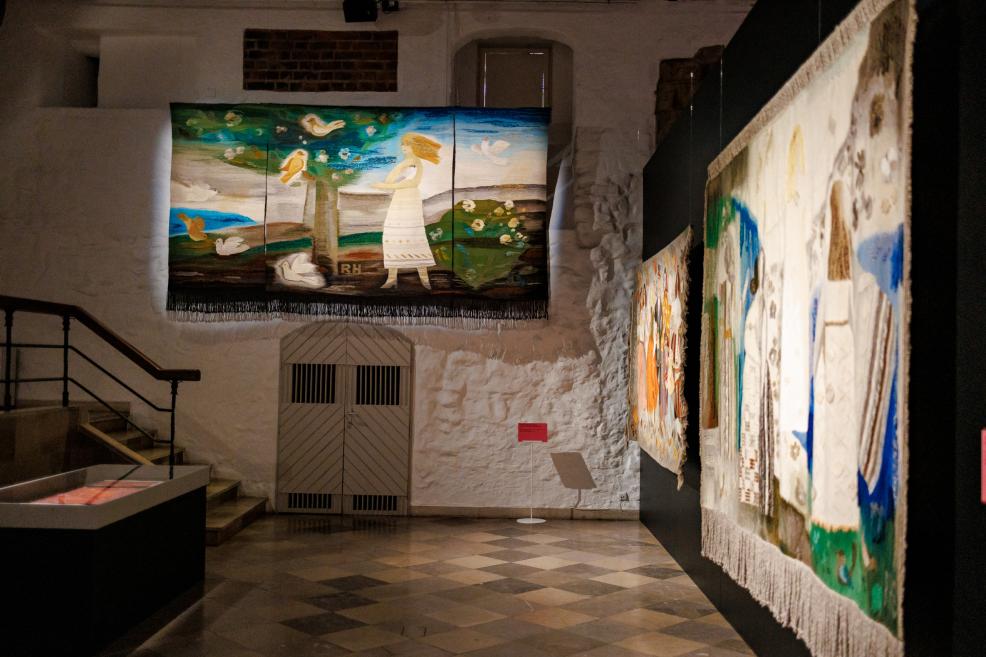Several large-format textile paintings hang on the walls in a museum gallery, including a central work depicting a woman and birds in a landscape.