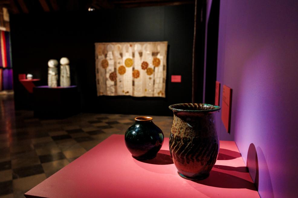 In an exhibition hall, two ceramic vases stand on a pink pedestal, with a textile work and other display pieces on the wall in the background.