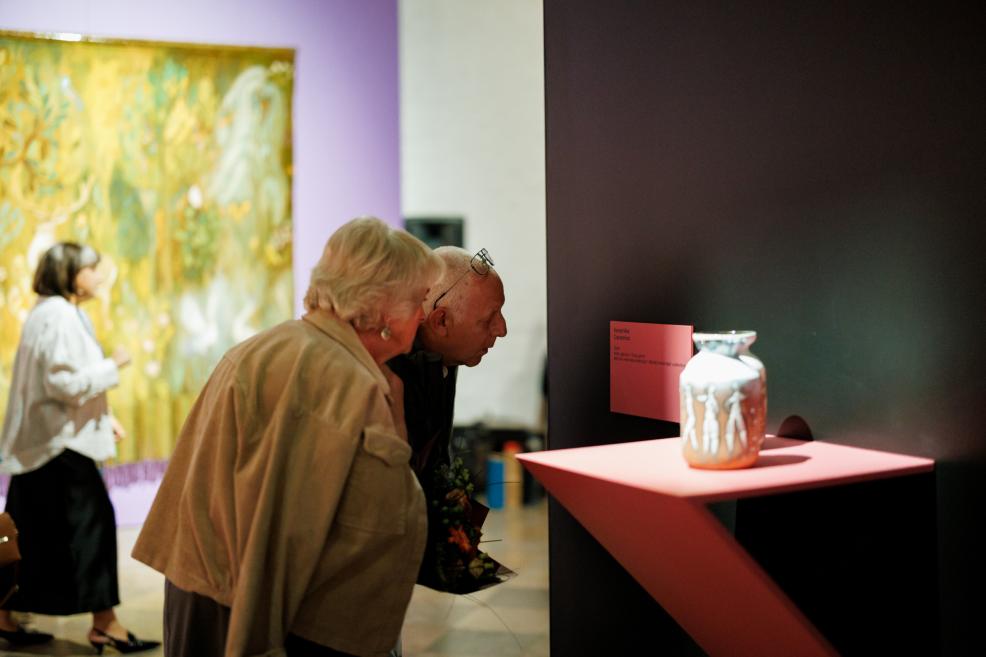 In a museum, two elderly visitors examine a ceramic vase displayed on a pink shelf against a dark wall, with a painting in the background.