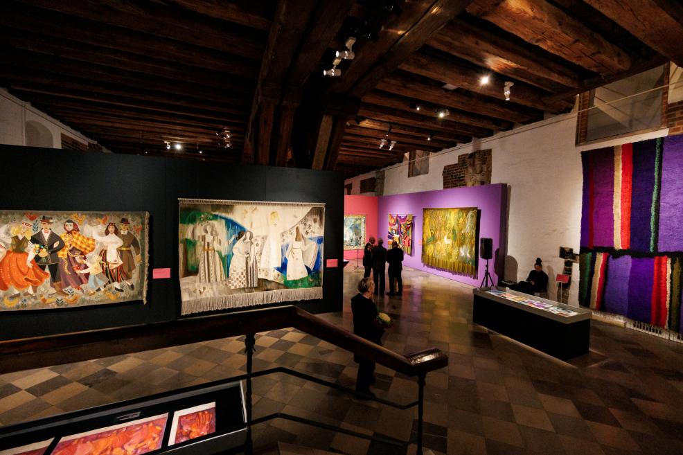 Large textile artworks and tapestries are displayed in a museum hall with wooden beams and a dark floor, with several visitors also visible in the room.