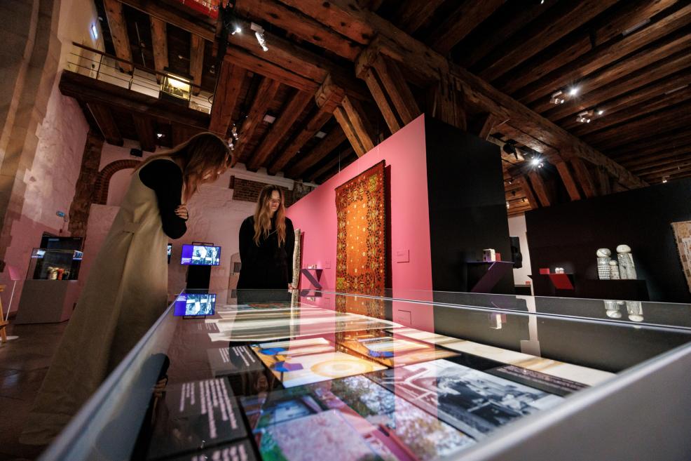 Museum exhibition hall with wooden ceiling beams, where two women view an exhibition with display cases, digital screens and a wall-mounted carpet.