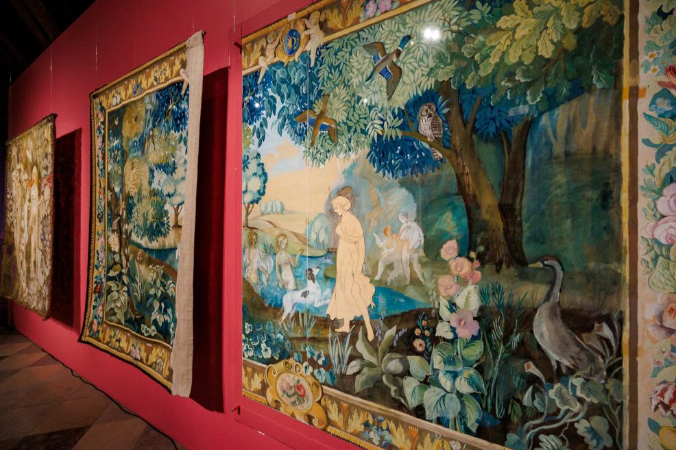 Large multicolored tapestry or textile compositions depicting a forest landscape with figures and birds displayed on a pink museum wall.
