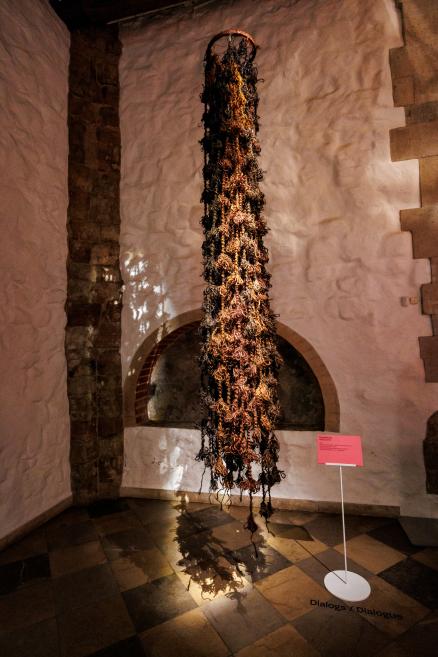 A long textile sculpture made of dark and reddish-brown threads hangs from the ceiling in a museum gallery with a wall niche and an information sign.