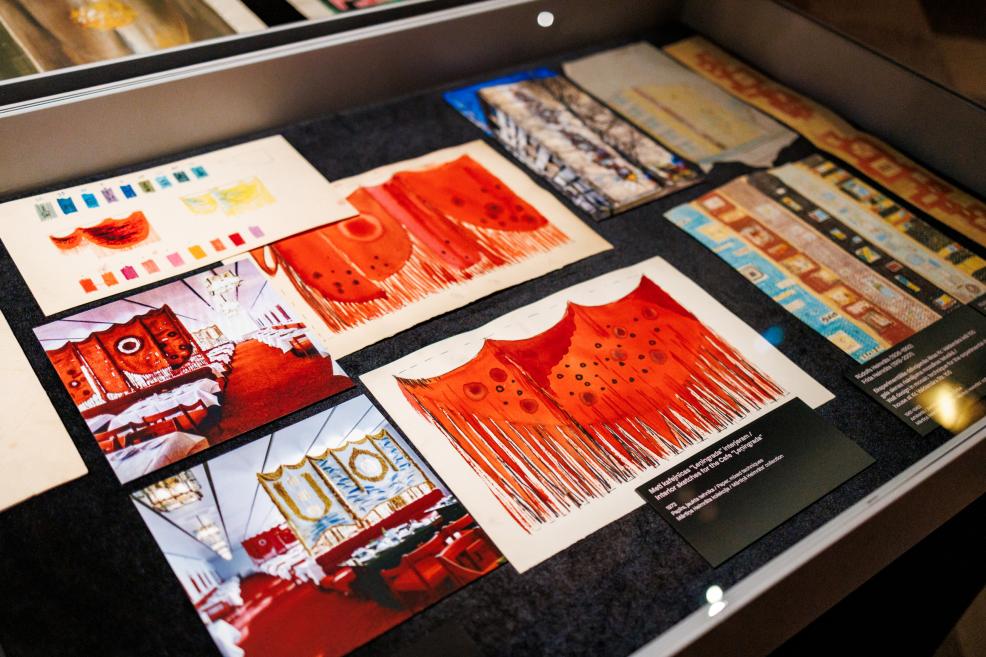 A display case contains several colorful textile and interior sketches, including red-brown and orange compositions, room visualizations, and pattern samples.