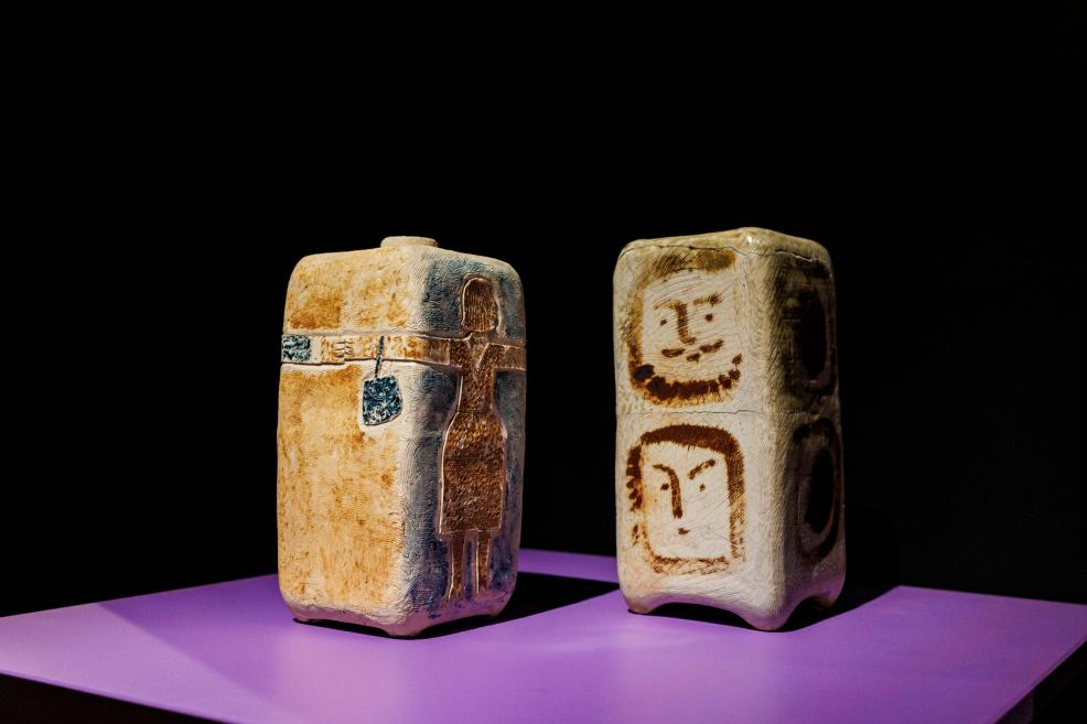Two vertical ceramic objects with stylized figures on a purple platform against a black background.