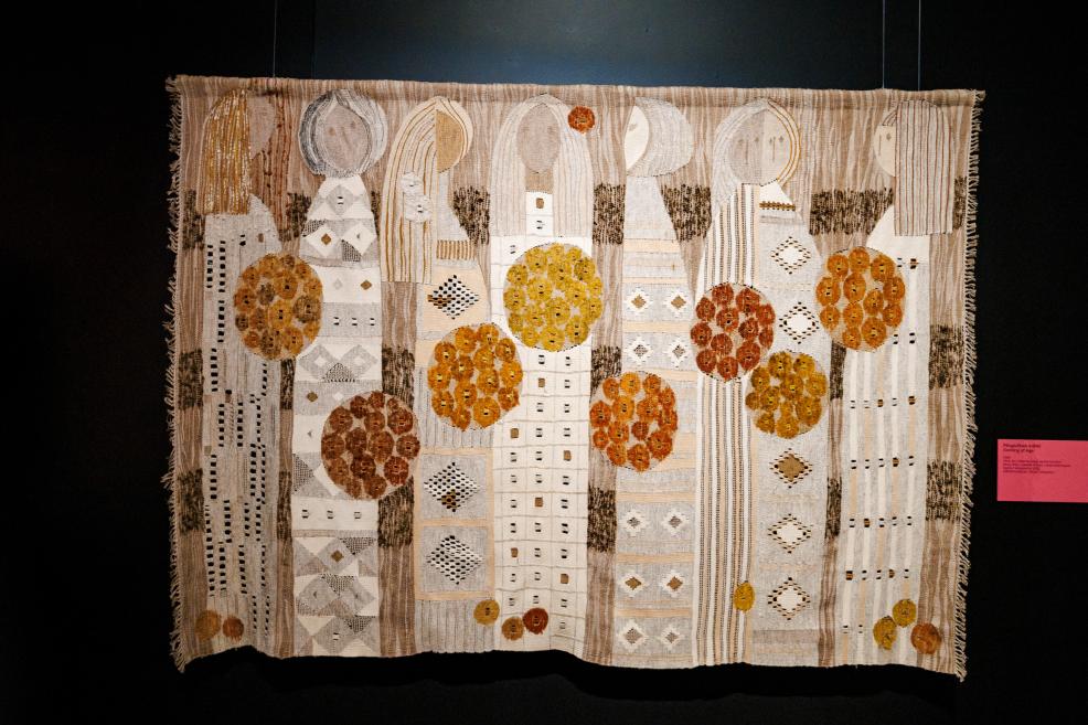 Textile tapestry with stylized tall figures, trees and geometric patterns in beige, white and ochre tones on a dark background.