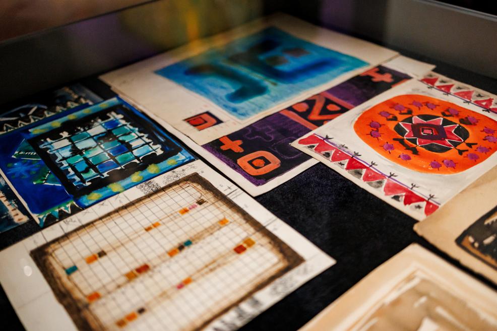 Several colorful abstract drawings on paper are arranged on a table, including blue, orange, and purple compositions with geometric patterns.