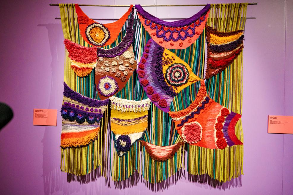 Colorful textile composition with hanging handmade relief forms and fringes on a purple museum wall.