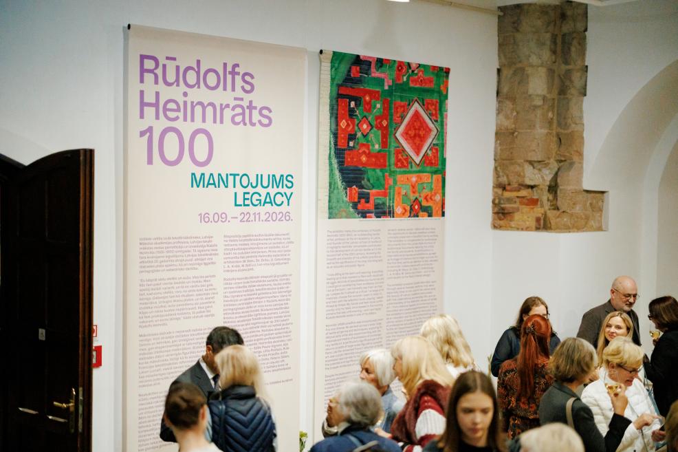 Izstādes zālē pie sienas redzami plakāti ar uzrakstu Rūdolfs Heimrāts 100 Mantojums Legacy un krāsaina tekstilmākslas darba reprodukcija, bet priekšplānā stāv vairāki apmeklētāji.
