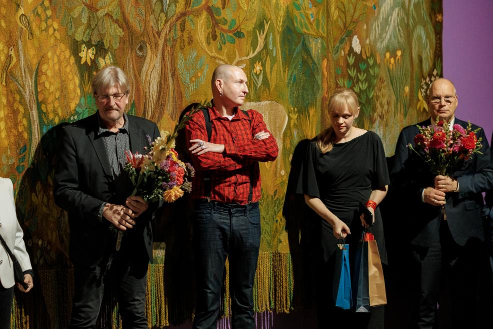 Četri cilvēki stāv pie dekoratīva gobelēna ar meža motīvu, divi no viņiem tur ziedu pušķus, bet sieviete rokās tur dāvanu maisiņus.