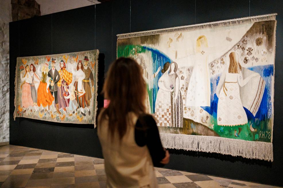 Muzeja zālē pie melnas sienas karājas divi lielformāta tekstila gobelēni ar cilvēku figūrām un ornamentālu rakstu, bet priekšplānā stāv skatītāja no muguras.