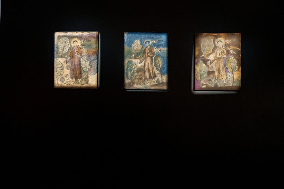 Trīs vertikāli gleznojumi ar stilizētu cilvēka figūru, kokiem un uzrakstiem, izvietoti uz tumša fona.