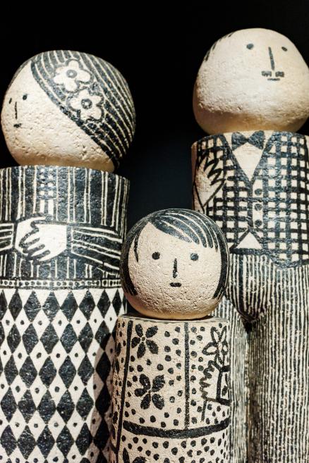 Dekoratīvu koka figūriņu grupa ar stilizētām sejām un melnbaltiem ģeometriskiem rakstiem uz tumša fona.