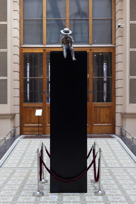 Muzeja interjerā uz augsta melna postamenta sēž mazs figūriņas veida skulpturāls cilvēks ar baltu cepuri, norobežots ar samta auklām.
