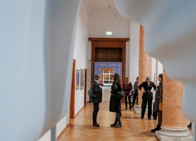 Muzeja izstāžu zālē redzamas uz baltiem paneļiem iekārtas glezniecības darbi, tostarp liela kompozīcija ar cilvēkiem pie galda un sievietes portrets.