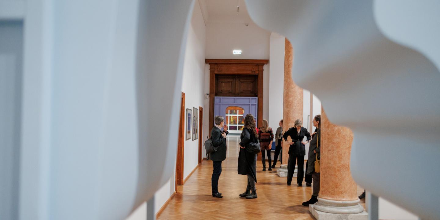 Muzeja izstāžu zālē redzamas uz baltiem paneļiem iekārtas glezniecības darbi, tostarp liela kompozīcija ar cilvēkiem pie galda un sievietes portrets.
