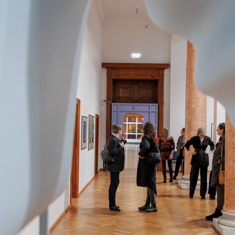 Muzeja izstāžu zālē redzamas uz baltiem paneļiem iekārtas glezniecības darbi, tostarp liela kompozīcija ar cilvēkiem pie galda un sievietes portrets.