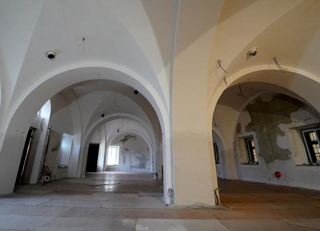Plaša telpa ar augstiem velvētiem griestiem, baltām sienām un logu ailēm, redzama renovācijas stadijā ar atklātiem vadiem un daļēji noņemtu apdari.