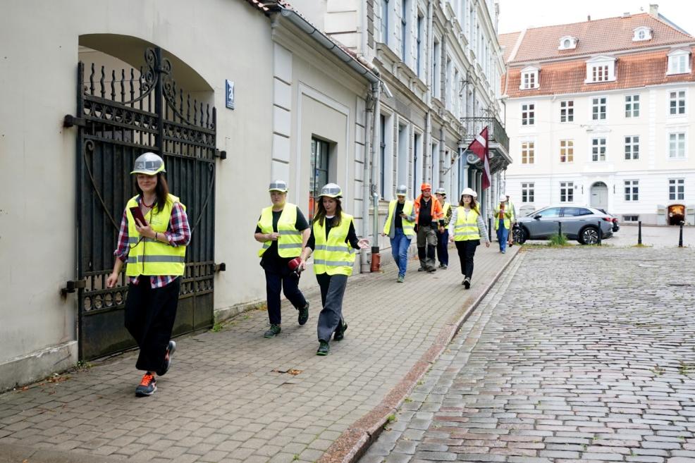 Aizsargķiverēs un atstarojošās vestēs tērpta grupa dodas pa bruģētu ielu gar vēsturisku ēku fasādēm.