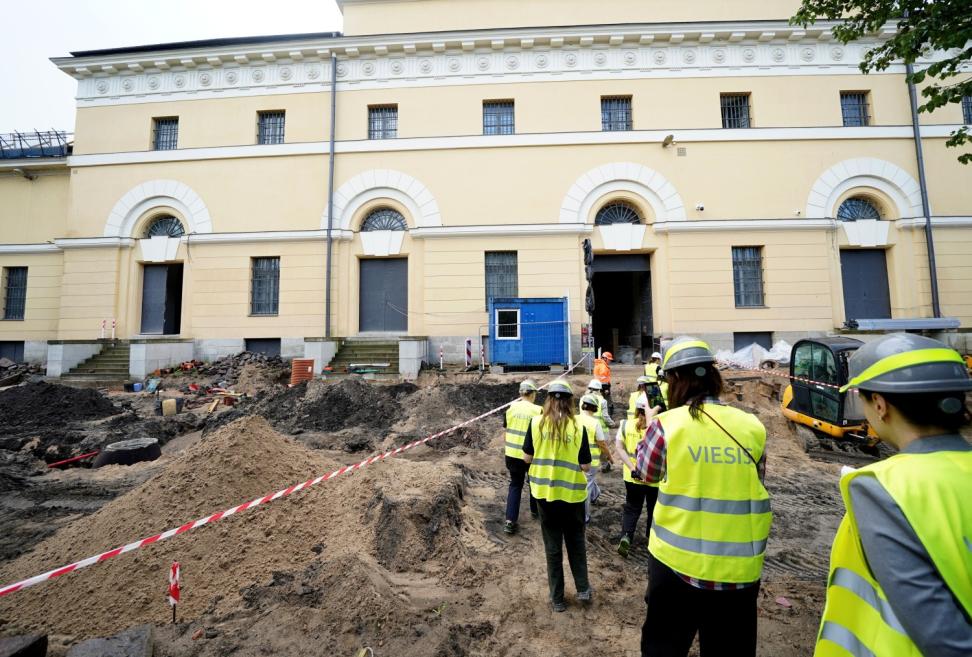 Darba drošības vestēs un ķiverēs tērpti cilvēki iet pa rakšanas vietu pie gaiši dzeltenas muzeja ēkas fasādes ar arkveida logiem un durvīm.