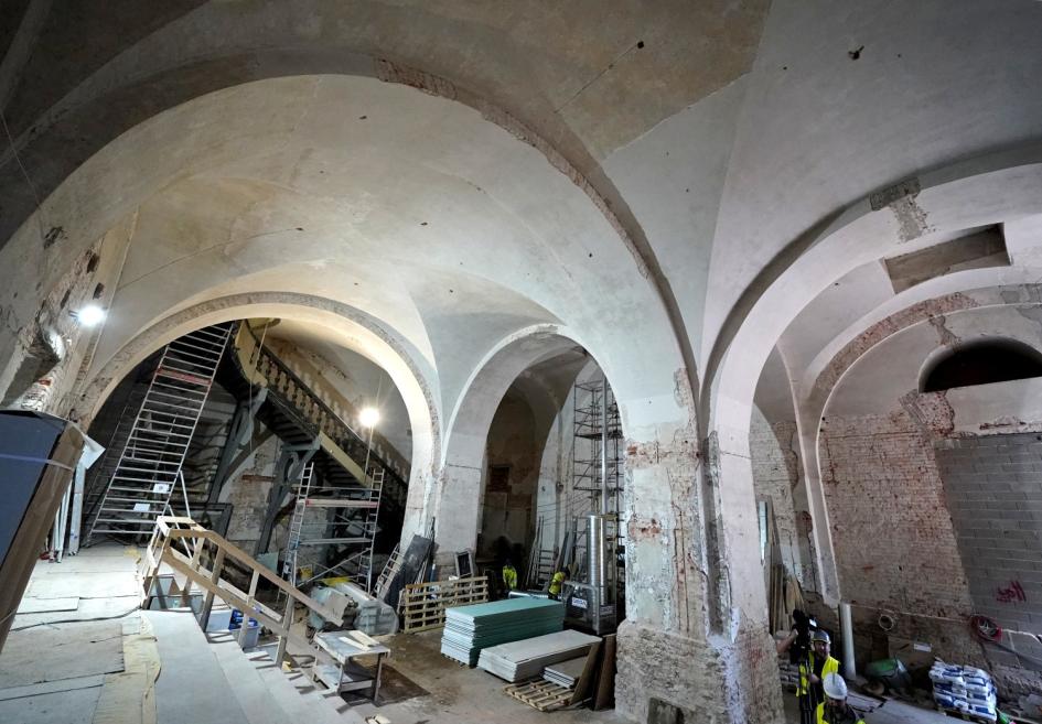 Plaša velvēta telpa ar būvdarbu stalažām, kāpnēm, materiālu kaudzēm un strādniekiem atsegto ķieģeļu sienu un apmetuma lokos.