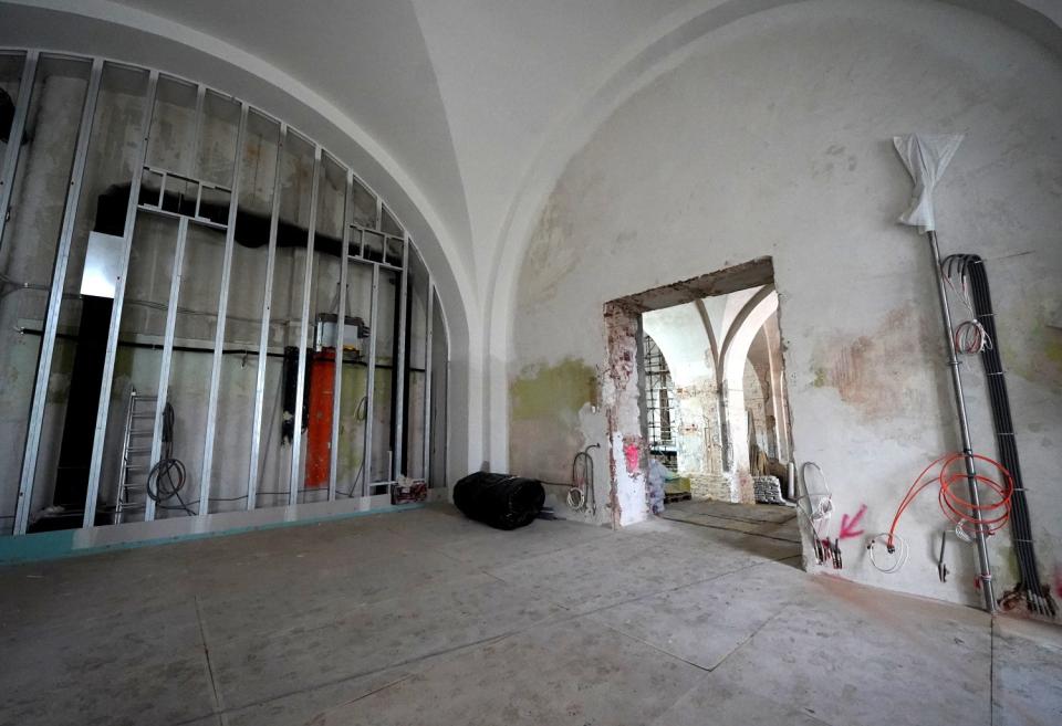 Restaurācijas stadijā esoša telpa ar velvju griestiem, atsegtiem metāla karkasiem, atvērtu durvju ailu un redzamiem elektrības vadiem pie sienām.