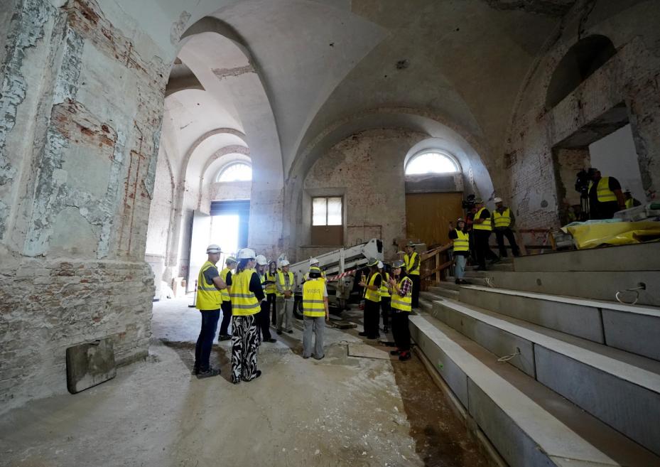 Arhitektoniska interjera telpa ar augstiem velvētiem griestiem, kurā vairākas personas dzeltenās drošības vestēs un ķiverēs apskata renovācijas darbus.
