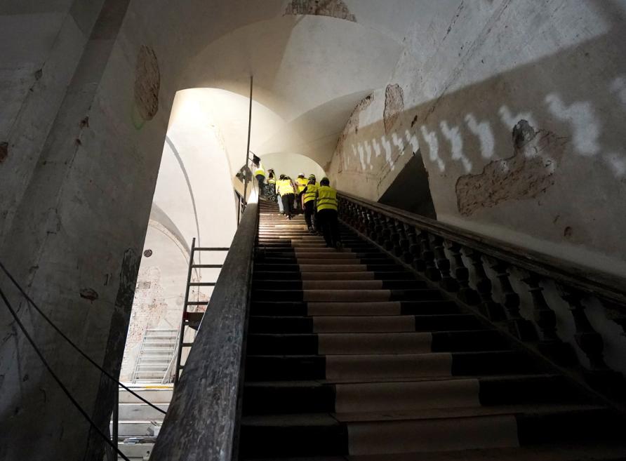 Vairāki celtnieki dzeltenās drošības vestēs kāpj pa stāvu kāpņu telpu ar velvētiem, daļēji bojātiem un remontējamiem griestiem.