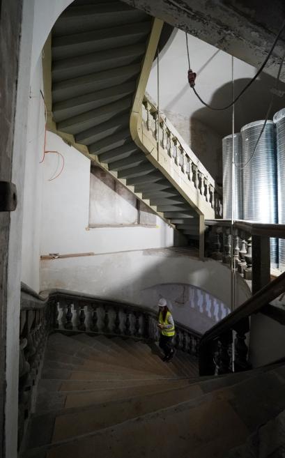 Vēsturiska spirālveida kāpņu telpa ēkas iekšpusē ar būvdarbu skatu: viena sieviete aizsargķiverē un atstarojošā vestē stāv uz kāpnēm pie margām.