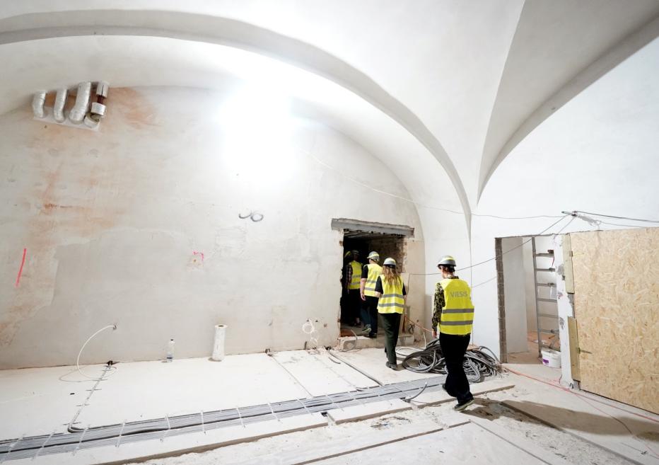 Renovācijas laikā redzama plaša velvēta telpa ar strādniekiem dzeltenās aizsargvestēs un ķiverēs, atvērumiem sienā un redzamiem kabeļiem uz grīdas.