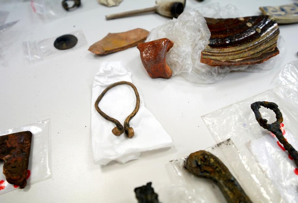 Uz balta galda izlikti un iepakojumos ievietoti arheoloģiski metāla un keramikas priekšmeti, tostarp rūsējušas detaļas, gredzens un salauzts keramikas fragments.