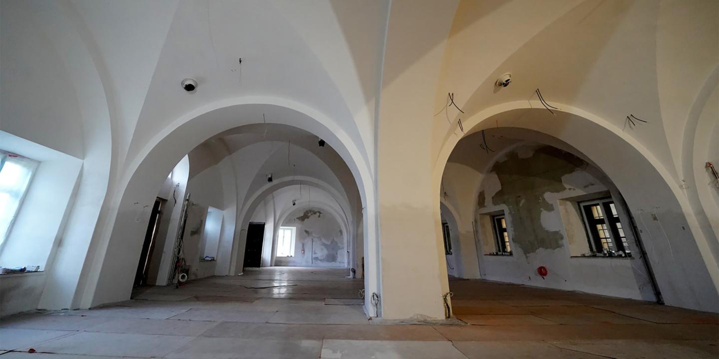 Plaša telpa ar augstiem velvētiem griestiem, baltām sienām un logu ailēm, redzama renovācijas stadijā ar atklātiem vadiem un daļēji noņemtu apdari.