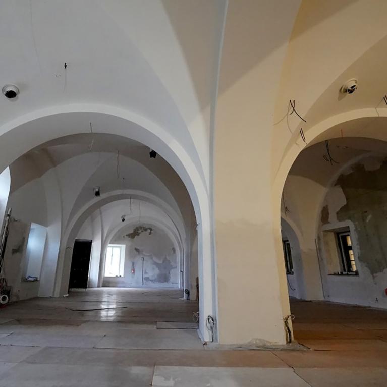 Plaša telpa ar augstiem velvētiem griestiem, baltām sienām un logu ailēm, redzama renovācijas stadijā ar atklātiem vadiem un daļēji noņemtu apdari.