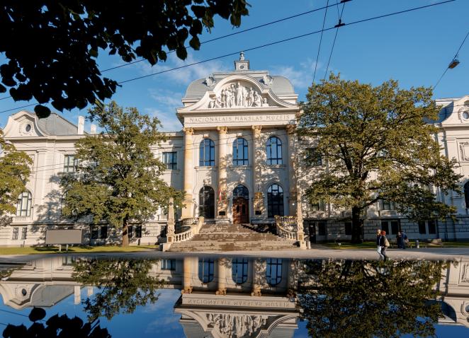 Latvijas Nacionālā mākslas muzeja vēsturiskā ēka redzama saulainā dienā, tās fasāde un apkārtējie koki atspīd ūdens virsmā ēkas priekšplānā.