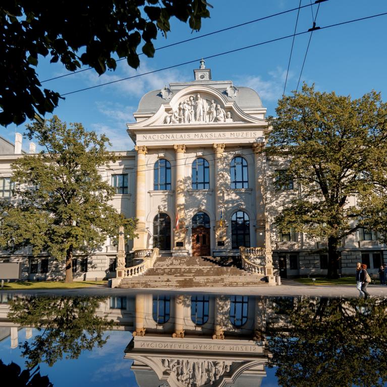 Latvijas Nacionālā mākslas muzeja vēsturiskā ēka redzama saulainā dienā, tās fasāde un apkārtējie koki atspīd ūdens virsmā ēkas priekšplānā.