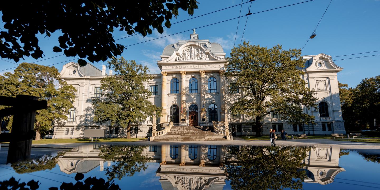 Latvijas Nacionālā mākslas muzeja vēsturiskā ēka redzama saulainā dienā, tās fasāde un apkārtējie koki atspīd ūdens virsmā ēkas priekšplānā.
