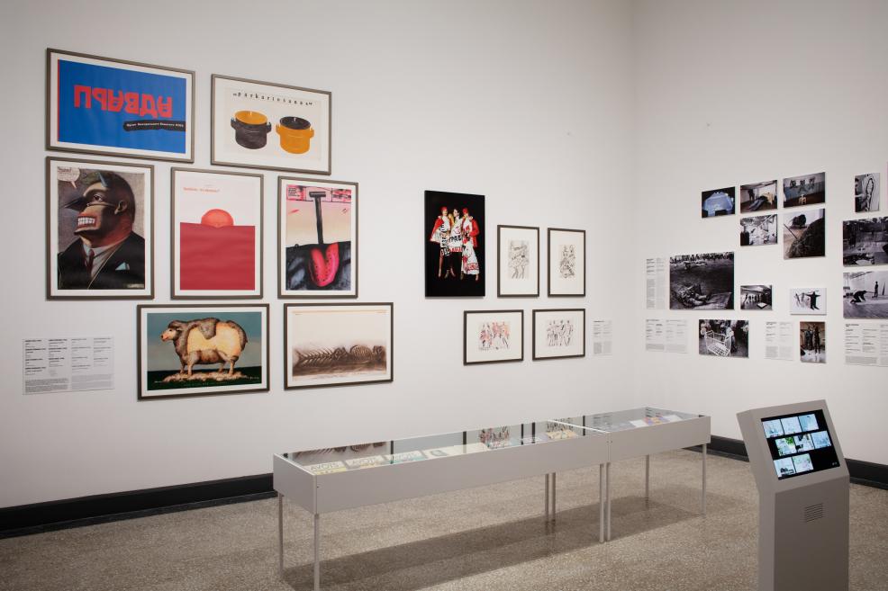 Muzeja ekspozīcijā uz baltām sienām izvietoti plakāti, grafikas darbi, fotogrāfijas un dokumentācijas materiāli, bet priekšplānā redzama vitrīna un digitāls ekrāns.