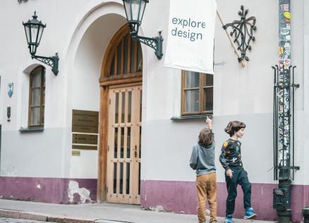 Divi bērni pie dizaina muzeja ēkas skatās uz karogu ar uzrakstu “explore design”.