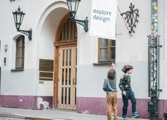 Divi bērni pie dizaina muzeja ēkas skatās uz karogu ar uzrakstu “explore design”.