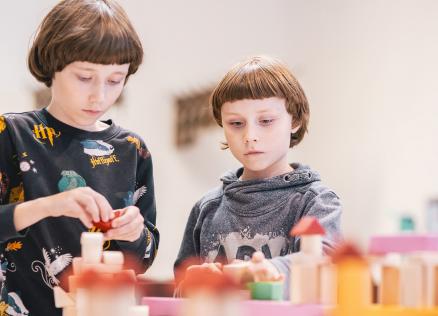 Divi bērni koncentrēti būvē konstrukcijas no krāsainiem koka klucīšiem gaišā telpā.