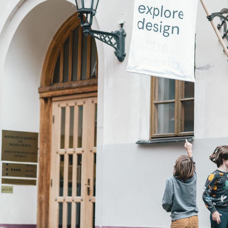 Divi bērni pie dizaina muzeja ēkas skatās uz karogu ar uzrakstu “explore design”.