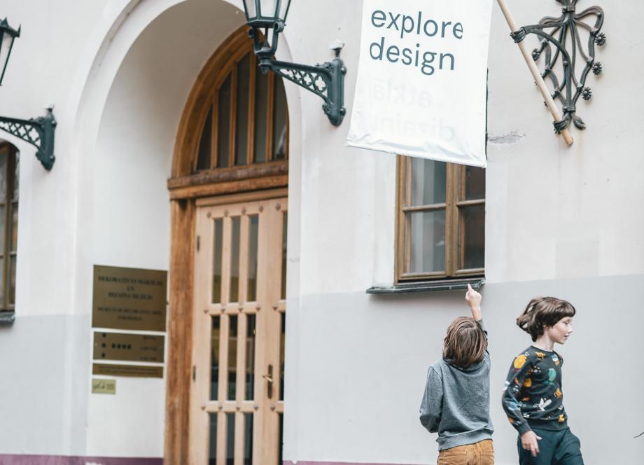 Divi bērni pie dizaina muzeja ēkas skatās uz karogu ar uzrakstu “explore design”.