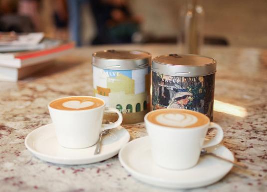 Divas kafijas tasītes ar sirds formas latte mākslu uz marmora galda. Aiz tām redzamas divas "Kalve" kafijas bundžas ar mākslinieciskiem dizainiem un uzrakstiem "Espresso blend of arts" un "Signature espresso blend. Toffee". Fonā – grāmatu kaudzīte un izpludināts interjera fons.