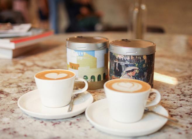Divas kafijas tasītes ar sirds formas latte mākslu uz marmora galda. Aiz tām redzamas divas "Kalve" kafijas bundžas ar mākslinieciskiem dizainiem un uzrakstiem "Espresso blend of arts" un "Signature espresso blend. Toffee". Fonā – grāmatu kaudzīte un izpludināts interjera fons.